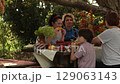Middle age woman cut pumpkin pie at Thanksgiving Day dinner. Diverse women with boy kid enjoying Thanksgiving dinner together, eating corn cobs at cozy autumn table under tree at fall garden party  129063143