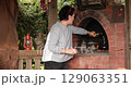 Woman add cooking oil into clay pot in rustic oven of fireplace. Outdoor cooking experience captures essence of traditional meal preparation in charming rustic environment. 129063351