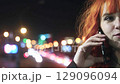 Beautiful red-haired woman smiling, talking on phone at night outdoors against city lights background 129096094