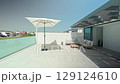 Sunlight illuminates a modern rooftop terrace, casting a shadow from a white parasol over the tiled floor, next to comfortable outdoor furniture 129124610