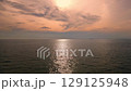 Aerial drone flight over the sea during sunset with distant orange-colored clouds and the sun setting on the horizon, creating a peaceful and warm tropical atmosphere.	 129125948