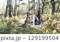 A cute young woman with a dog enjoys the beauty of nature and communication with pets. The woman drinks tea from a thermos during a picnic in the autumn forest 129199504
