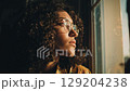 Thoughtful woman with curly hair, glasses, looking out sunlit window. 129204238