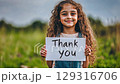 Young girl expresses gratitude outdoors while holding a sign in a green field 129316706