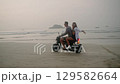 Young people drive motorbike on sea beach at sunset. Man, woman travel by motorcycle on coastline along ocean. Female hugs boyfriend, raises arms to air, touches hair blown in the wind. Guy rides bike 129582664
