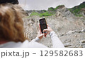 Woman hiker takes photos and videos of mountains. She explores nature and documents rocky landscape with smartphone. Outdoor adventure on sunny day. Capturing travel memories. Slow motion. 129582683
