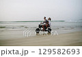 Young couple ride motorcycle, splashing water on wet sandy sea beach at sunset. Man, woman have fun driving by motorbike on ocean coast. Lady raises hand to air, hugs boyfriend on romantic trip. 129582693