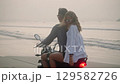 Young couple riding motorcycle on ocean beach at sunset. Man, woman travelling by motor bike on coastline along sea. Lady looks back to camera, laughs, spreads arms, touches hair blowing in the wind. 129582726