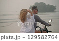 Couple in sunglasses ride motorcycle on sea beach at sunset. Lady in swimwear hugs guy, touches wind blown hair, smiles at camera on romantic bike drive. Man, woman travel on motorcycle by ocean. 129582764