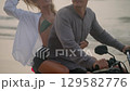 Elegant couple ride motorcycle along sea beach on sunrise. Intraframe installation from bike to people. Tourists drive rented motorbike on ocean coast. Woman hugs man, spreads arms riding scooter. 129582776