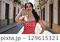 Dressed in a striking red dress adorned with polka dots, model strolls through charming streets on a summer travel adventure. Captivating local culture and stunning architecture surround her. 129615321