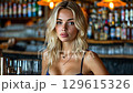 In a lively bar, a blonde woman stands proudly in front of a well stocked shelf of spirits, exuding charm and confidence while dressed in a fashionable outfit suited for a night out with friends. 129615326