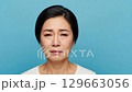 A sorrowful Asian female adult is shown in a headshot, crying with a deeply distressed expression and wet cheeks, isolated on a vibrant blue studio backdrop. 129663056