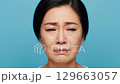 A sorrowful Asian female is depicted in a front-facing close-up, with tears streaming down her cheeks, expressing profound grief on a bright blue studio backdrop. 129663057