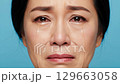 An extreme close-up features an East Asian woman's face as she weeps, with visible tears flowing, expressing a strong emotion of sorrow or grief on a teal studio backdrop. 129663058