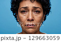 The emotional face of a mature woman with dark skin and curly hair is captured in a headshot, showing tears and red eyes, conveying profound grief on a bright blue studio backdrop. 129663059