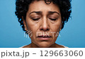 A front-facing, headshot perspective shows an emotional individual with a sorrowful expression and multiple tears visibly flowing down their cheeks, set against a plain blue studio backdrop. 129663060