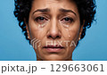 An emotional close-up portrays a mature woman with dark, curly hair experiencing deep sadness, with clear tears running down her cheeks against a solid blue backdrop. 129663061