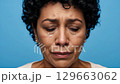 A front-facing view of a sorrowful woman with dark, curly hair, her face wet with tears and eyes reddened from crying, isolated on a vibrant blue studio backdrop. 129663062