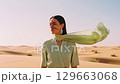 A beautiful young female model poses in a sandy desert, wearing an elegant mint green outfit with a long scarf dramatically blowing in the wind, showcasing fashion and nature. 129663068