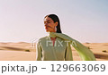 A young adult woman with dark hair and elegant earrings, dressed in a light green pleated garment with a flowing scarf, stands looking away across sun-drenched sand dunes. 129663069