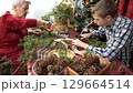 Grandmother and grandson make Christmas wreath together. Elderly woman and teenage boy glue pine branches 129664514