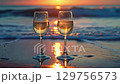 Two glasses of sparkling wine illuminated by the setting sun. Two glasses filled with sparkling wine stand on the wet sand, gently washed by the waves, sparkling in the rays of the sunset. 129756573