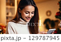 Young woman in white sweater smiling while holding a smartphone near a festive Christmas tree with golden lights. zoom in, zoom out, pan, or 360 degree shot, animate 129787945