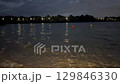 Evening view of a calm lake with small waves, orange and yellow buoys floating on the water. Shoreline in the foreground, city lights and trees in the distance. Nighttime mood, peaceful nature concept 129846330