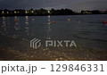 Evening view of a calm lake with small waves, orange and yellow buoys floating on the water. Shoreline in the foreground, city lights and trees in the distance. Nighttime mood, peaceful nature concept 129846331