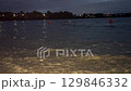 Evening view of a calm lake with small waves, orange and yellow buoys floating on the water. Shoreline in the foreground, city lights and trees in the distance. Nighttime mood, peaceful nature concept 129846332