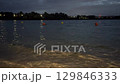 Evening view of a calm lake with small waves, orange and yellow buoys floating on the water. Shoreline in the foreground, city lights and trees in the distance. Nighttime mood, peaceful nature concept 129846333