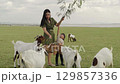 mother with toddler girl feeding a tree branch to the goat on grass field near the river 129857336
