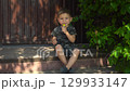 Three year old boy with a bruised knee sucking a lollipop sitting on the porch during a hot summer day in dark clothes 129933147