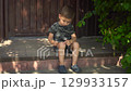 The child looks closely at his scraped knee. He gently touches the scab with his finger. He is sitting on a wooden porch on a hot summer day. He is wearing a t-shirt, shorts and slippers. 129933157