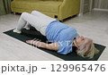 Elderly woman performing glute bridge exercise on yoga mat 129965476