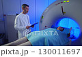 Medical worker standing confidently near MRI tunnel while composed patient lying flat on scanning table. Operating diagnostic system during scheduled medical procedure inside clean examination area. 130011697