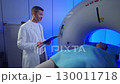 Medical worker standing confidently near MRI tunnel while composed patient lying flat on scanning table. Operating diagnostic system during scheduled medical procedure inside clean examination area. 130011718