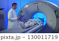 Medical worker standing confidently near MRI tunnel while female patient resting on table. Patient lies on MRI table and undergoes magnetic resonance imaging. Man in robe and protective cap enters 130011719