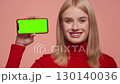 Smiling woman holding smartphone with green screen in horizontal position on pink background 130140036