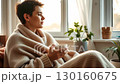 Young woman with a mug sits by the window wrapped in a blanket. Cozy atmosphere of warmth, comfort, and peaceful home relaxation. Generative AI Looping video. 130160675
