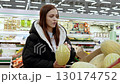 Young woman inspecting a ripe melon in a supermarket's fruit section, carefully evaluating fresh produce while shopping for nutritious food choices 130174752