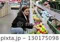 Young woman crouching in a grocery aisle, selecting vibrant bell peppers from the shelf and placing them into a plastic bag. Fresh produce fills the surrounding space 130175098
