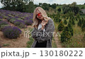 Blonde flirty smile woman wearing casual attire standing in lavender field, holding vibrant red poppy near eye, enjoying scenic rural landscape. Playful smile of blonde woman holding red poppy flower 130180222