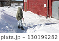 Moving snow with manual snow shovel by individual dressed in winter clothing standing beside large snow pile near rural house. 130199282