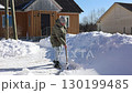 Shoveling snow with wooden handled snow shovel on rural property with person wearing warm jacket hat and boots near house. 130199485