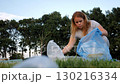 A young female volunteer, wearing gloves, collects plastic garbage in a park, against the backdrop of a parking lot, as part of an effort to protect the environment. 130216334