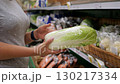 Close-up of a woman selecting fresh Peking cabbage from a large supermarket. Shelves full of vegetables and fruits in the grocery aisle. 130217334