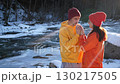A caring and loving man holds the hands of his beloved wife as they stand near a mountain river on a cold winter day. They wearing bright sweatshirts, on the background of the winter forest and river. 130217505