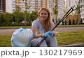 A tired but happy female volunteer is sitting on the curb, holding a full garbage bag in her hands. The activists have cleared the park area of plastic garbage as part of the urban development project 130217969